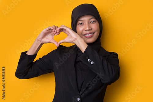 Portrait asian muslim woman wearing hijab showing heart sign isolated on yellow background
