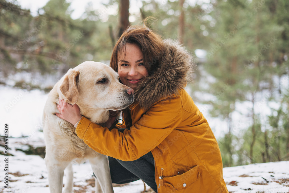 Young smiling woman in yellow jacket with big kind white dog Labrador walking in winter forest