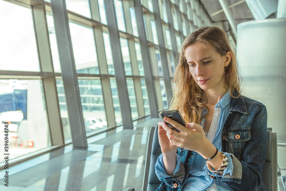 Portrait of young beautiful long-haired woman looking and typing on phone with message and sitting on background of panoramic window of modern airport building.