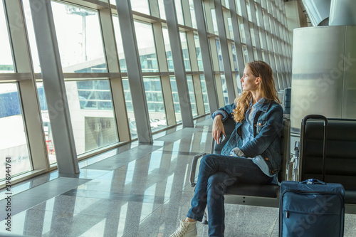 Portrait of a young long-haired woman looking pensively out of the window and holding a phone in her hand and sitting indoor in the waiting room on background of a panoramic window of a airport.