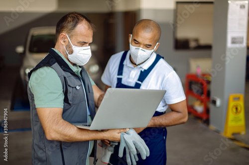 Two auto mechanics wearing face masks while working on laptop in a workshop.