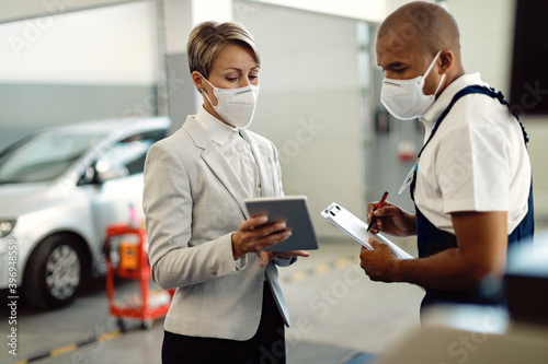 Businesswoman and African American mechanic wearing face masks and cooperating while using touchpad in a workshop.