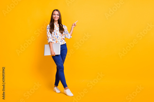 Photo portrait full body view of girl pointing finger at blank space holding laptop in one hand isolated on vivid yellow colored background