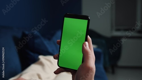 Man lying on couch using smart phone with chroma key green screen at night, scrolling through social media or online shop - internet, communications concept close up.