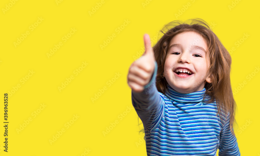 A little girl of Caucasian appearance in a blue striped turtleneck shows a thumbs up and laughs, smiling happily. Yellow background, copy space