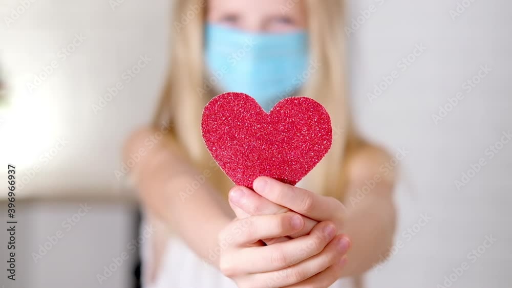 Red heart close up in hands of little blonde girl in medical mask on Valentines Day. Safety Covid holiday celebration. Slow motion Red heart close up in hands of little blonde girl in medical mask on Valentines Day. Safety Covid holiday celebration. Slow motion