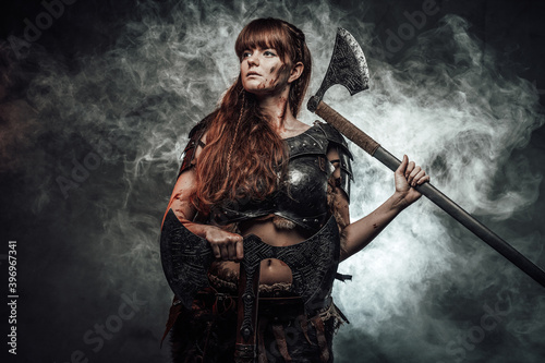 Barbaric female viking in light armour with brown hairs poses in dark smokey background holding two axes.