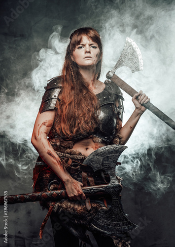 Armed with two huge axes scandinavian female warrior with brown hairs in dark armour poses in smokey background. © Fxquadro