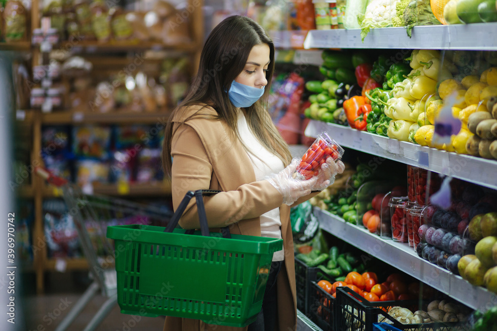 Happy woman in face mask taking fresh cherry tomatoes while standing at groceries in supermarket. Beautiful young girl with food basket choosing red tomatoes by stand with vegetables. Quarantine