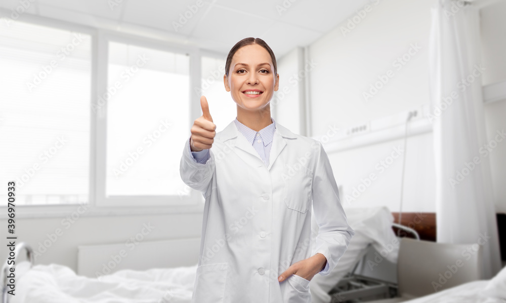 medicine, profession and healthcare concept - happy smiling female doctor in white coat showing thumbs up over hospital ward background