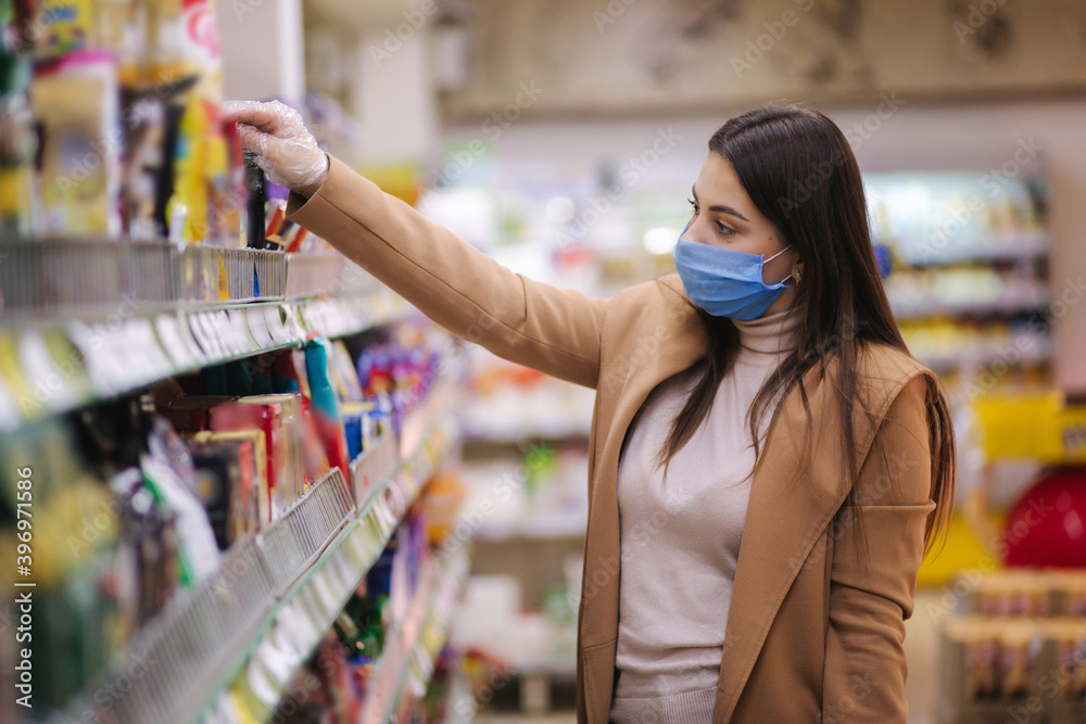 Horizontal shot of woman in protective mask and gloves reading label with price at goods while standing in supermarket. Beautiful young girl choosing snaks while doing shopping at grocery shop