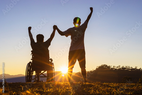 Vászonkép Silhouette of joyful disabled man in wheelchair raised hands with friend at suns