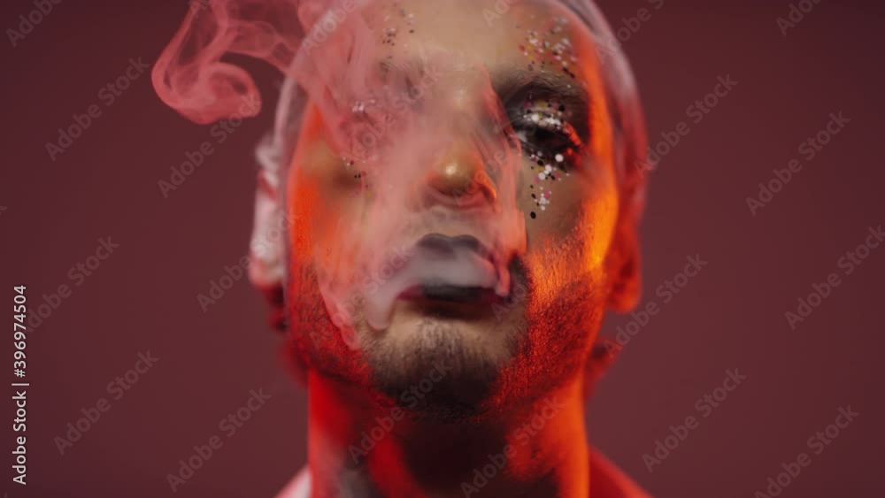 Portrait shot of young man with dramatic make-up and glitter blowing smoke and looking at camera in studio with red lighting Portrait shot of young man with dramatic make-up and glitter blowing smoke and looking at camera in studio with red lighting