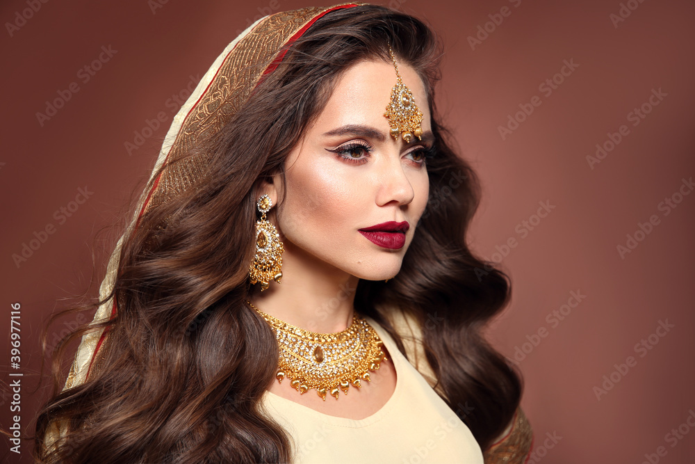  Hermosa mujer con peinado largo ondulado y maquillaje brillante.  Modelo morena hindú joven con conjunto de joyas de oro kundan aislado sobre fondo de estudio marrón.  foto de Stock