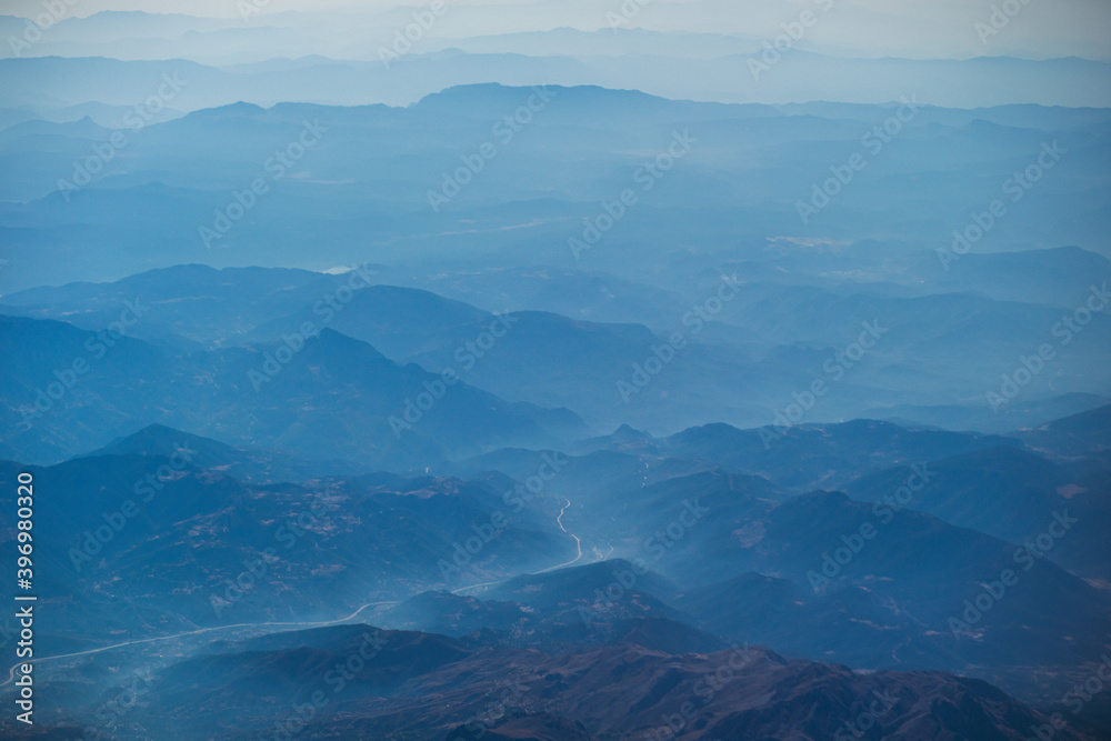 Scenic view at foggy landscape from flying plane. View from window at beautiful morning soft white transparent clouds and silhouettes of mountains.