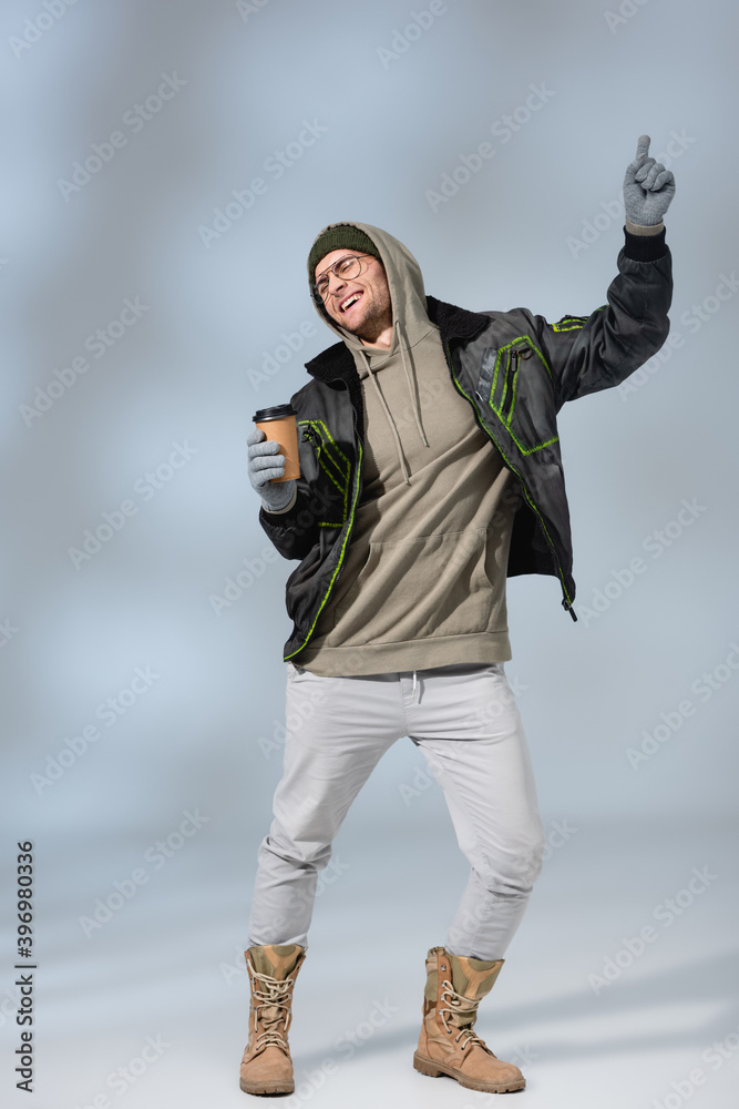 Φωτογραφία full length of young happy man in hat, hoodie and anorak holding paper cup and d