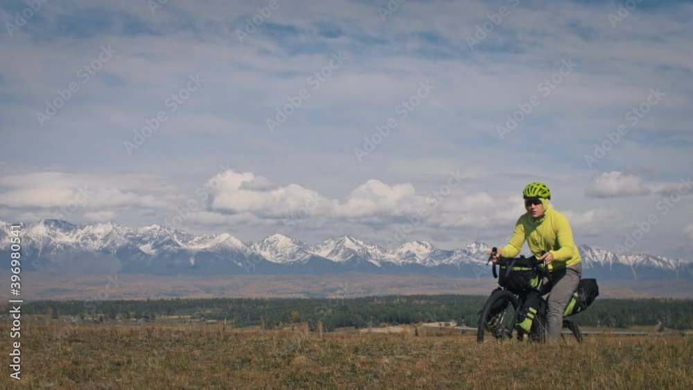 The man travel on mixed terrain cycle touring with bikepacking. The traveler journey with bicycle bags. Sport tourism bikepacking, bike, sportswear in green black colors. Mountain snow capped. The man travel on mixed terrain cycle touring with bikepacking. The traveler journey with bicycle bags. Sport tourism bikepacking, bike, sportswear in green black colors. Mountain snow capped.