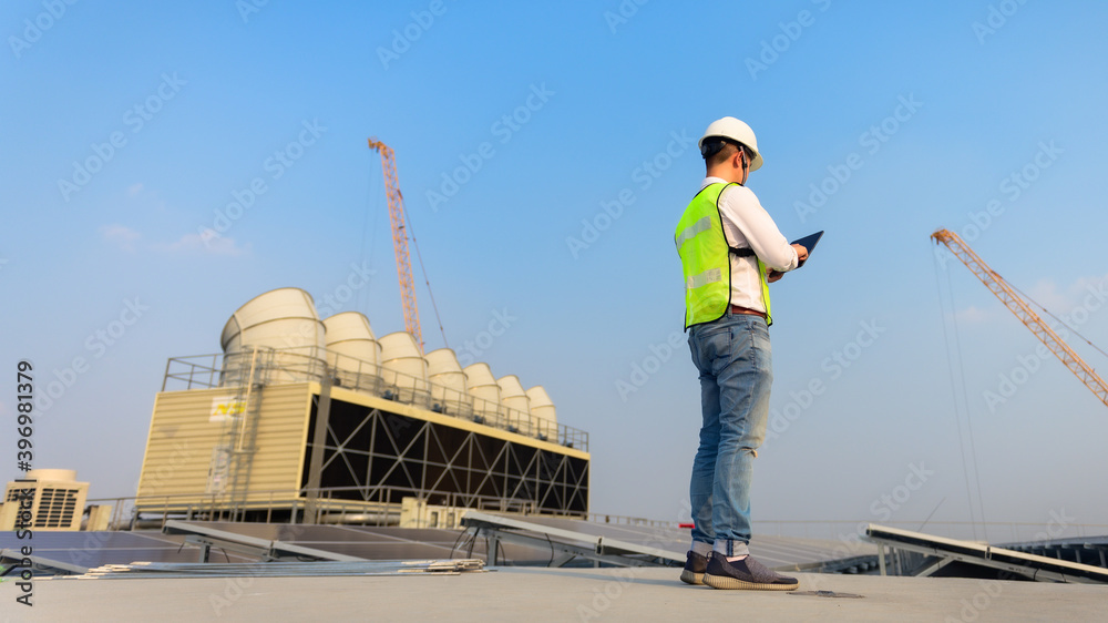 Engineer holding tablet is checking the cooling tower and solar cell on the roof of the building to be in good condition.