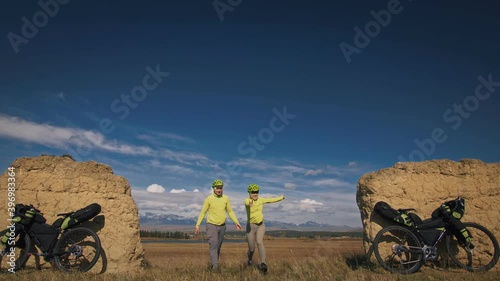 The man and woman travel on mixed terrain cycle bike touring with bikepacking. The two people journey with bicycle bags. Sport sportswear in green black colors. Mountain snow capped, stone arch.
