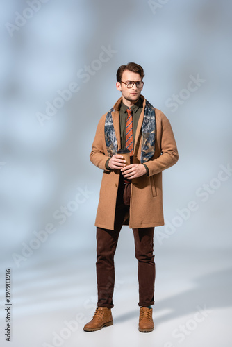 full length of stylish man in winter coat holding paper cup on grey photo