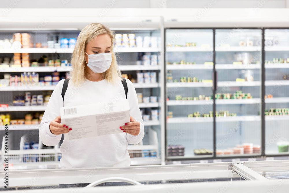 Young blond woman in a medical mask in a supermarket in the frozen food department. Coronavirus pandemic.