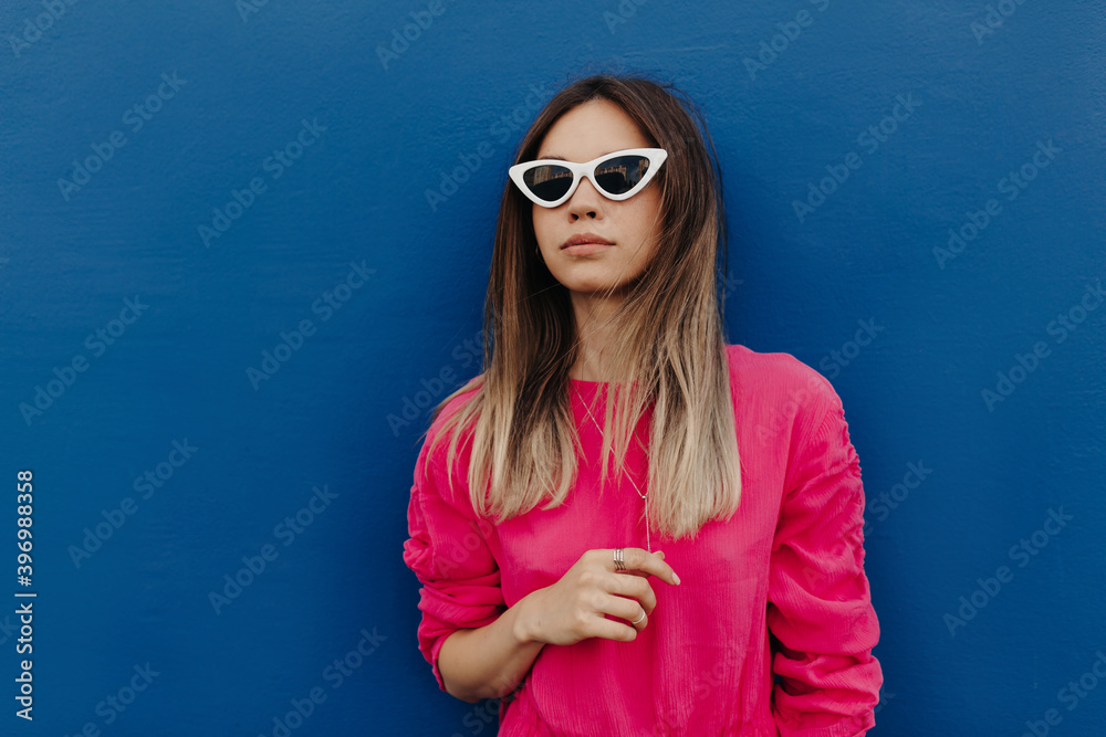 Close up outdoor portrait of charming young woman wearing pink shirt and white sunglasses posing over isolated blue wall Close up outdoor portrait of charming young woman wearing pink shirt and white sunglasses posing over isolated blue wall