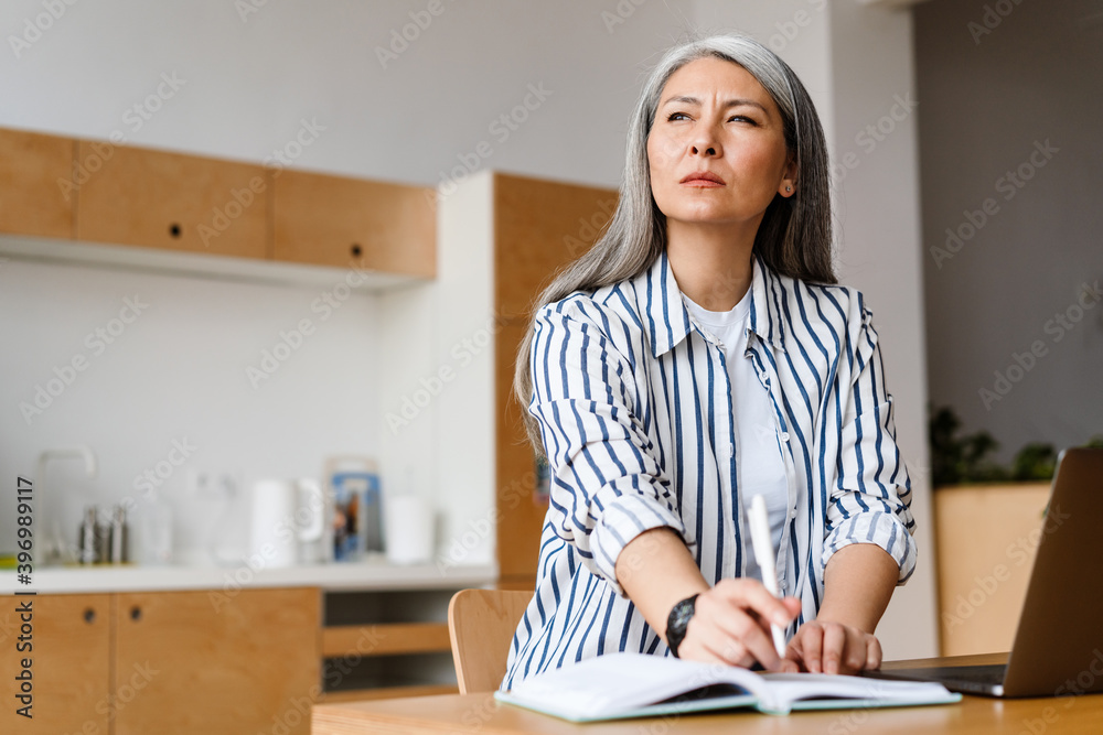 Serious white-haired mature woman writing down notes working with laptop Serious white-haired mature woman writing down notes working with laptop