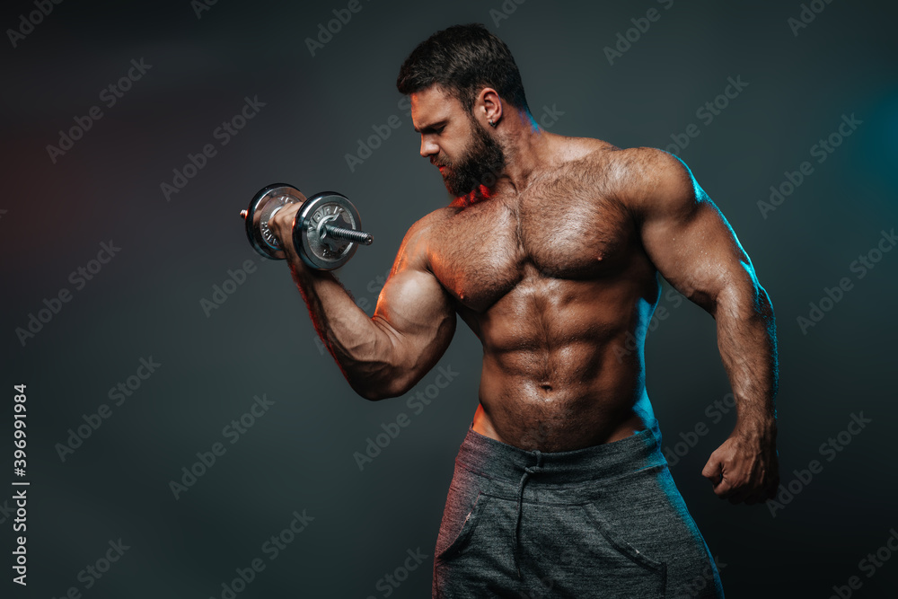 Muscled shirtless man with dumbbell doing his biceps exercise. Male body in colourful lightning in studio. Big hunk with six pack abs exercises in studio at grey background.
