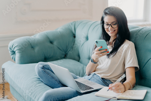 Skilled female student makes presentation for project, searches necessary information via mobile phone, has opened laptop on knees, wears casual clothes, poses on sofa in cozy room, chats online