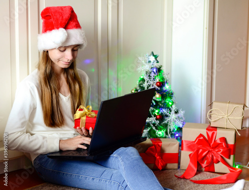 Girl with long hair and blue eyes in a Santa hat communicates online, wishes Christmas. Concept of online shopping, gifts, greetings.