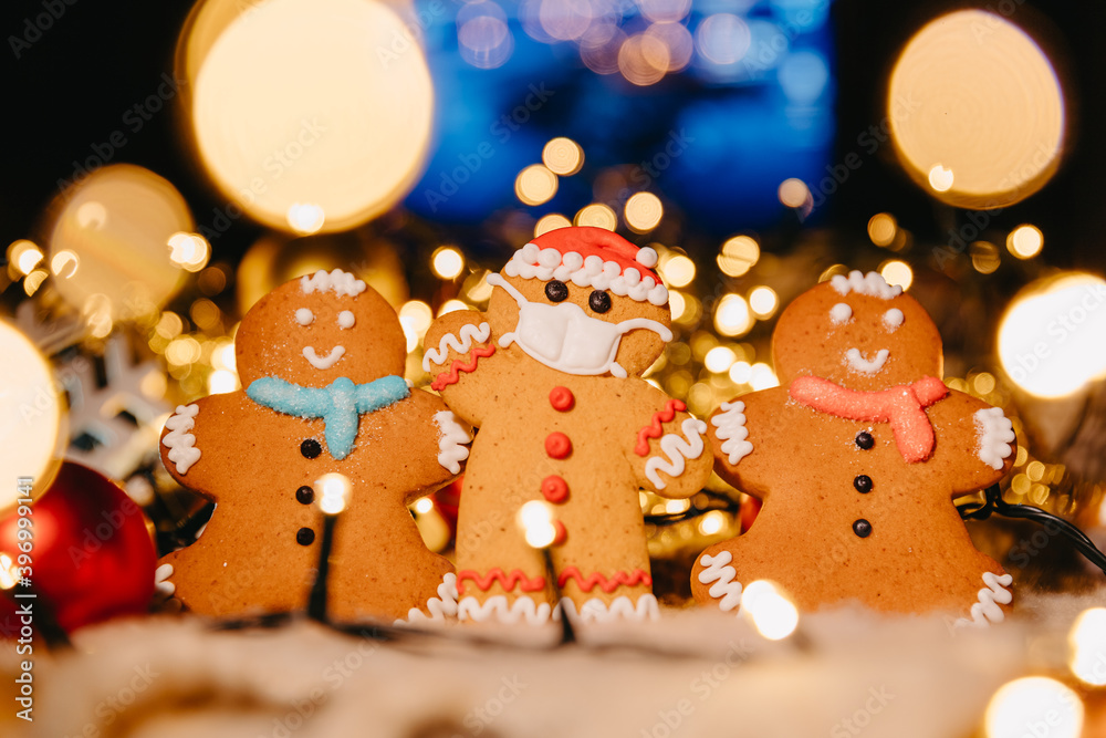Coronavirus pandemic, conceptual background with unfocused lights christmas decorations and gingerbread in the shape of Santa Claus wearing a protective medical mask