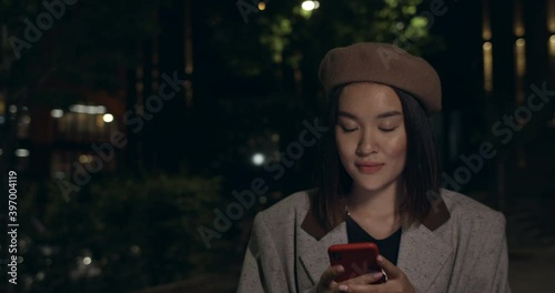 Attractive woman using smartphone and than looking to camera while walking at street. Stylish female person in beret chatting in social media. Concept of communicationand night life. photo