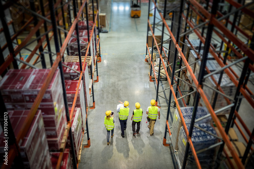 Quadro em tela Warehouse manager and workers consult plans walking between tall shelves, above