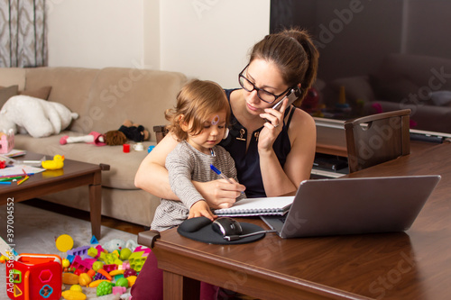 Young beautiful business woman talking on mobile phone and working on a laptop. Mother playing with child during working. Women powerful. Lady working at home during quarantine because of coronavirus.