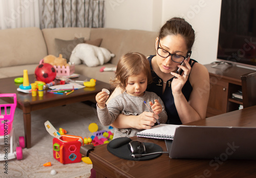 Young beautiful business woman talking on mobile phone and working on a laptop. Mother playing with child during working. Women powerful. Lady working at home during quarantine because of coronavirus.