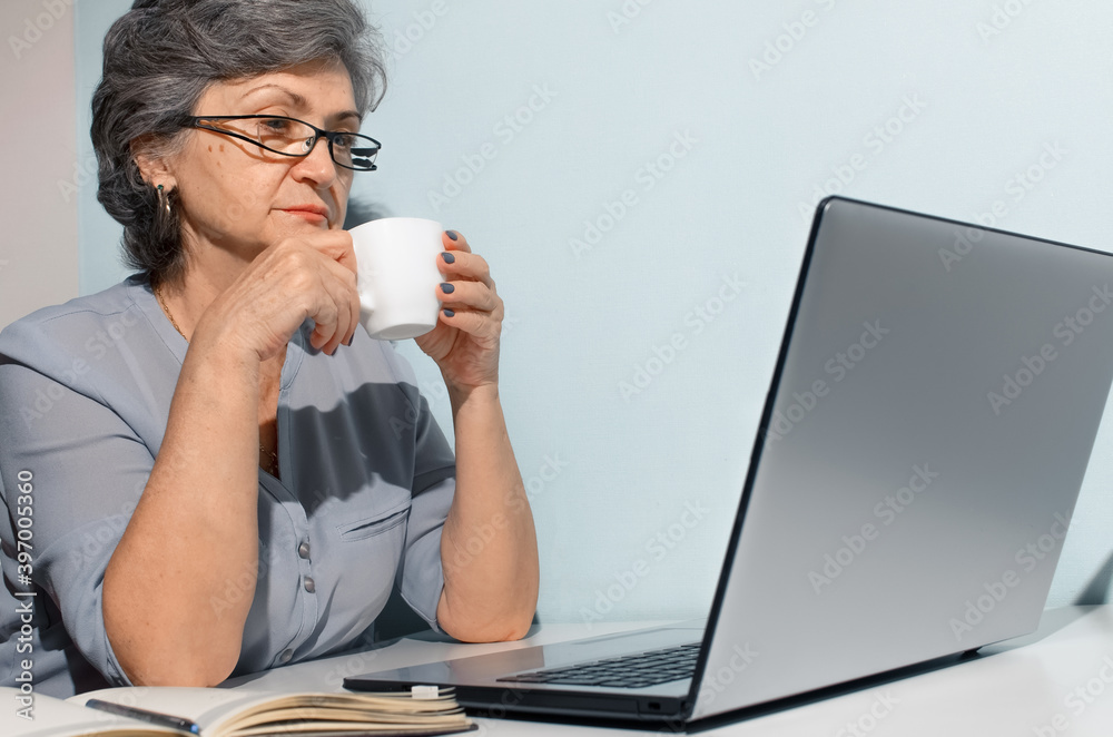 Senior woman with glasses studying online at home. Elderly woman drinking coffee at laptop. Concept of telecommuting, freelancing, retired work Senior woman with glasses studying online at home. Elderly woman drinking coffee at laptop. Concept of telecommuting, freelancing, retired work