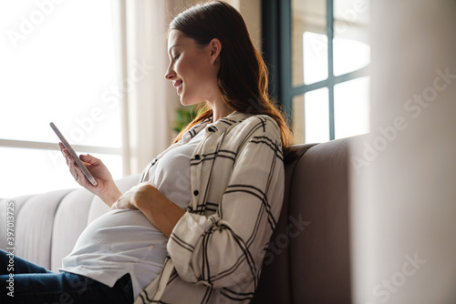 Happy charming pregnant woman using mobile phone while sitting on sofa photo