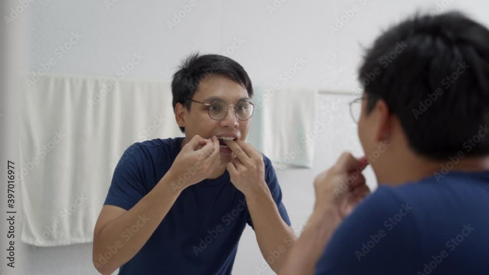 asian man using dental floss removing food particles that left on teeth off in mirror at white bathroom. asian man using dental floss removing food particles that left on teeth off in mirror at white bathroom.