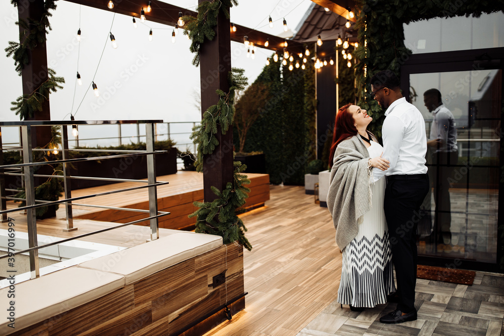 Happy couple stand at the balcony, handsome African American man hug gorgeous white woman wrapped in a blanket, enjoy tender moments, winter holidays, Christmas morning concept