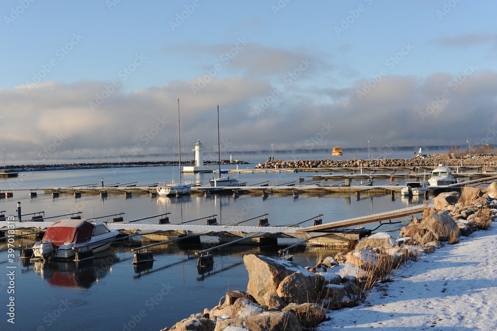 A pier on a harbour on Vättern lake in Gränna in Sweden on a sunny winter  day with white snow, blue sky, clear air and water, a lighthouse and yachts  Stock Photo