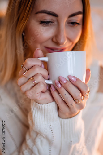 Portrait of a pretty blonde in a cozy home sweater with a Cup of tea or coffee in her hands with rings and a beautiful manicure.