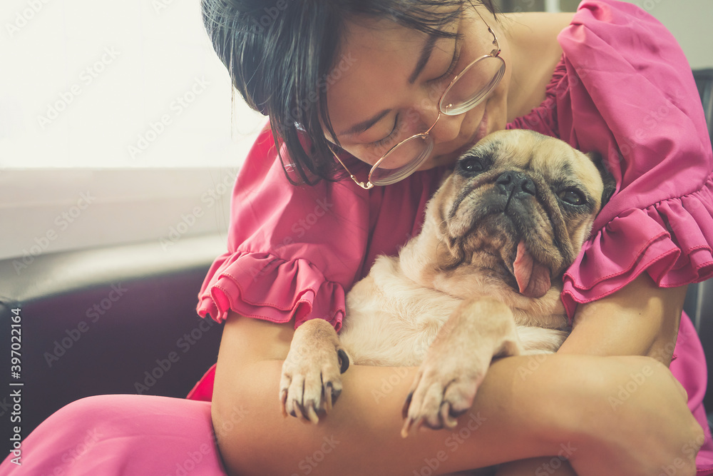 Beautiful young asian woman sitting on a sofa kissing and cuddling a cute pug dog.Young woman playing with her dog at home.