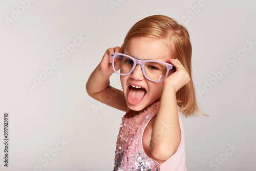 Kid girl shows her tongue. Posing in pink glasses and dress over grey background with copy space