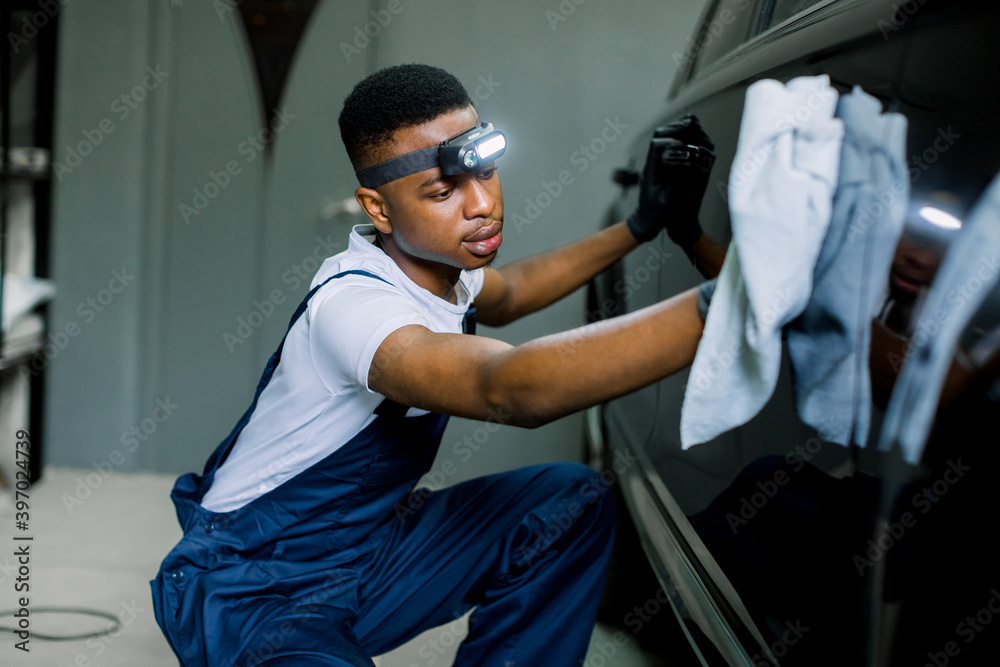 Young black car wash worker man, wearing protective clothes and gloves, cleaning modern black car with microfiber cloth. Car detailing or valeting concept. Selective focus on face. Young black car wash worker man, wearing protective clothes and gloves, cleaning modern black car with microfiber cloth. Car detailing or valeting concept. Selective focus on face.