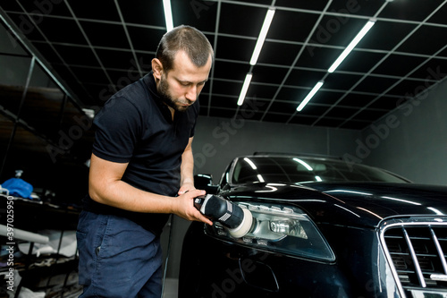 Car detailing and polishing concept. Close up of male worker with orbital polisher, polishing headlight of black luxury car at auto repair service © sofiko14