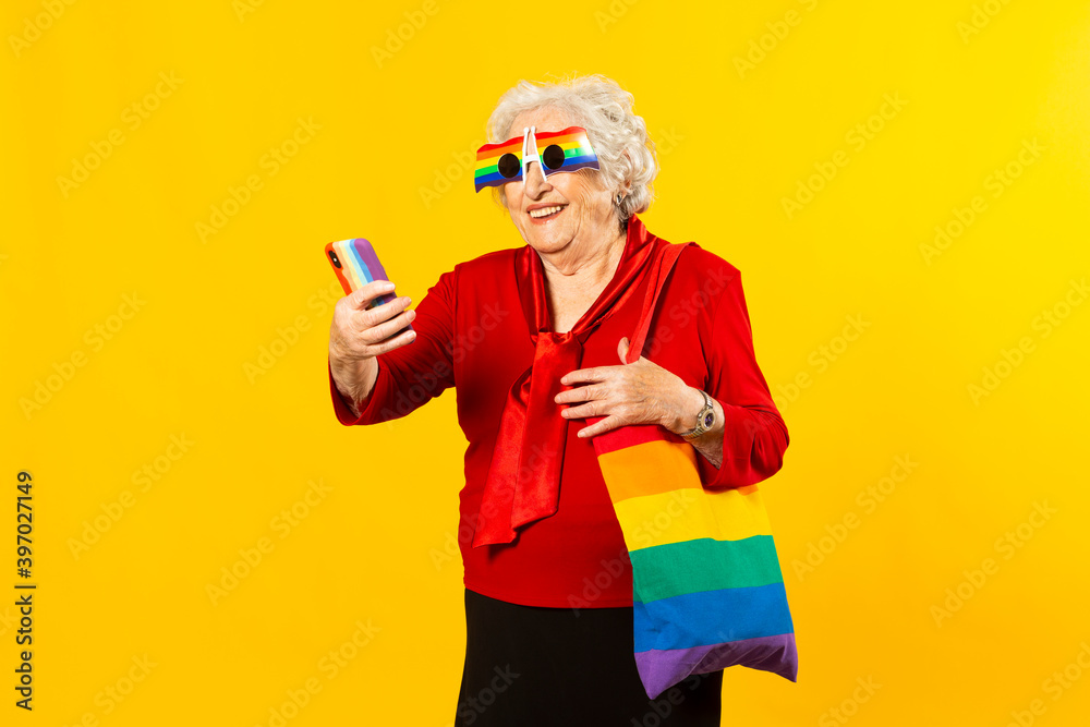 Studio portrait of a senior woman wearing a red shirt, rainbow sunglasses and a bag, having a video call with her mobile phone, against a yellow background Studio portrait of a senior woman wearing a red shirt, rainbow sunglasses and a bag, having a video call with her mobile phone, against a yellow background