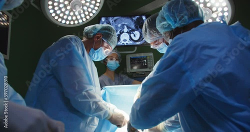 Group of mixed-races professional surgeons and nurses in uniform performing heart transplant surgery operation under bright lamps using medical instruments in operating room looking at screen photo