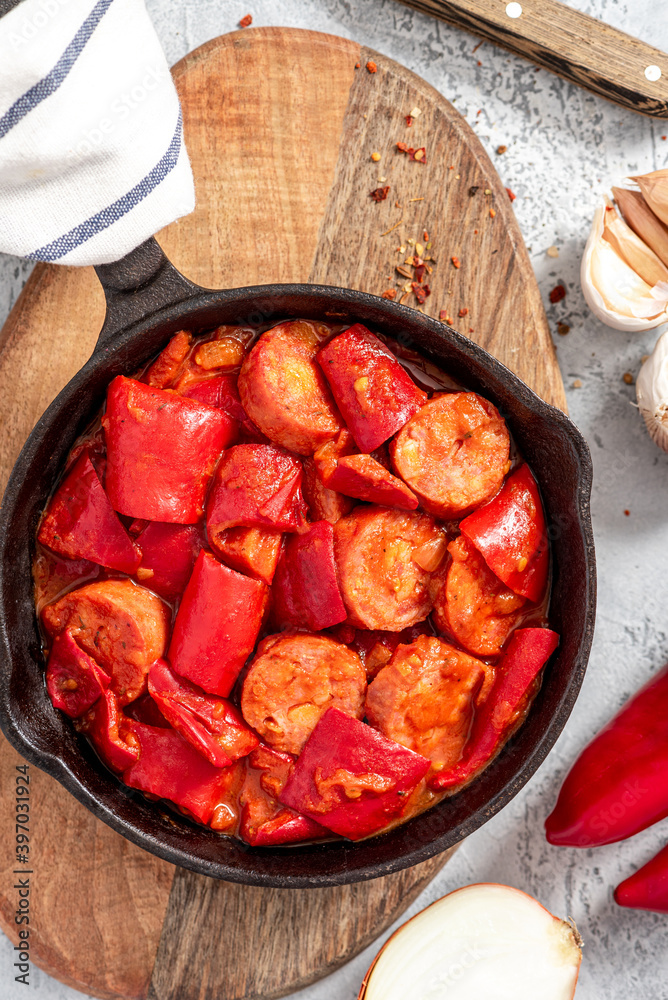 Sausages stewed with peppers, onions and tomatoes in a pan on a gray concrete background. Traditional Hungarian food lecho with sausages and vegetables.