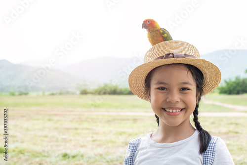 Asian small cute child is playing a colourful small bird, bird is standing on hat. Cute smiling girl playing with her pet green Sun Conure parrot. Exotic pet.