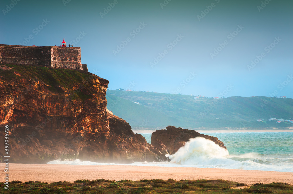 Big waves on the coast of Atlantic ocean in Nazare, Portugal. View of the cape with lighthouse from the North beach